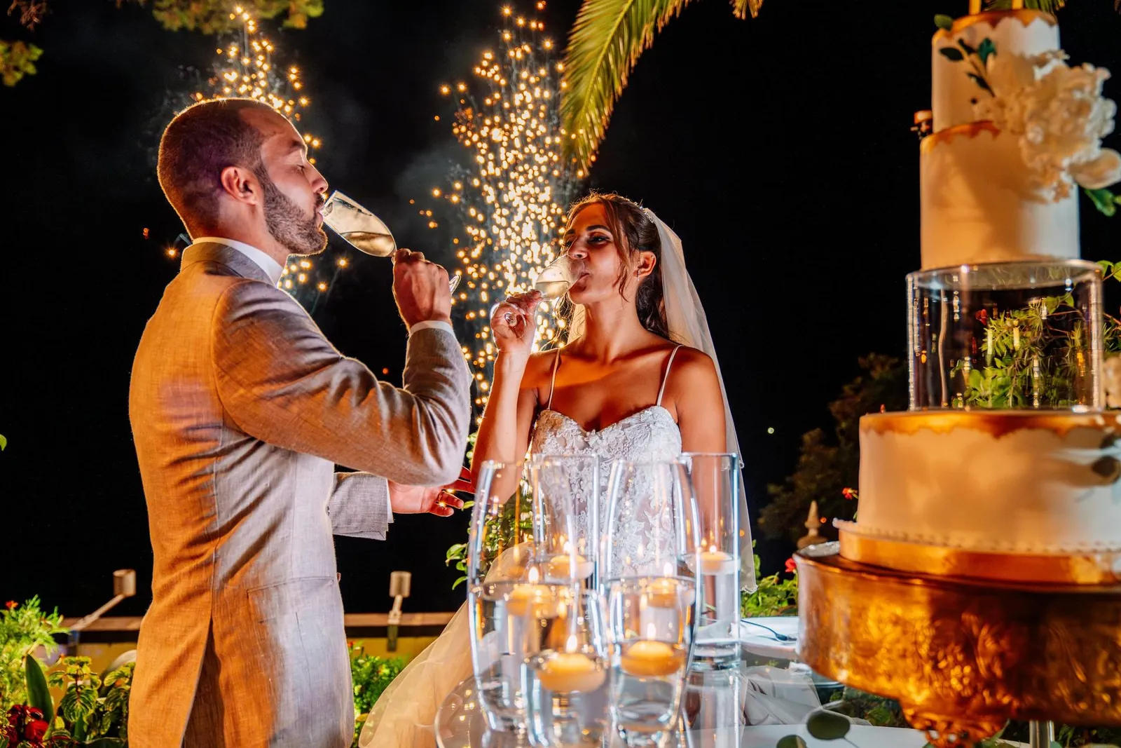 Noivos brindam junto ao bolo, à noite, com o fogo de artifício atrás