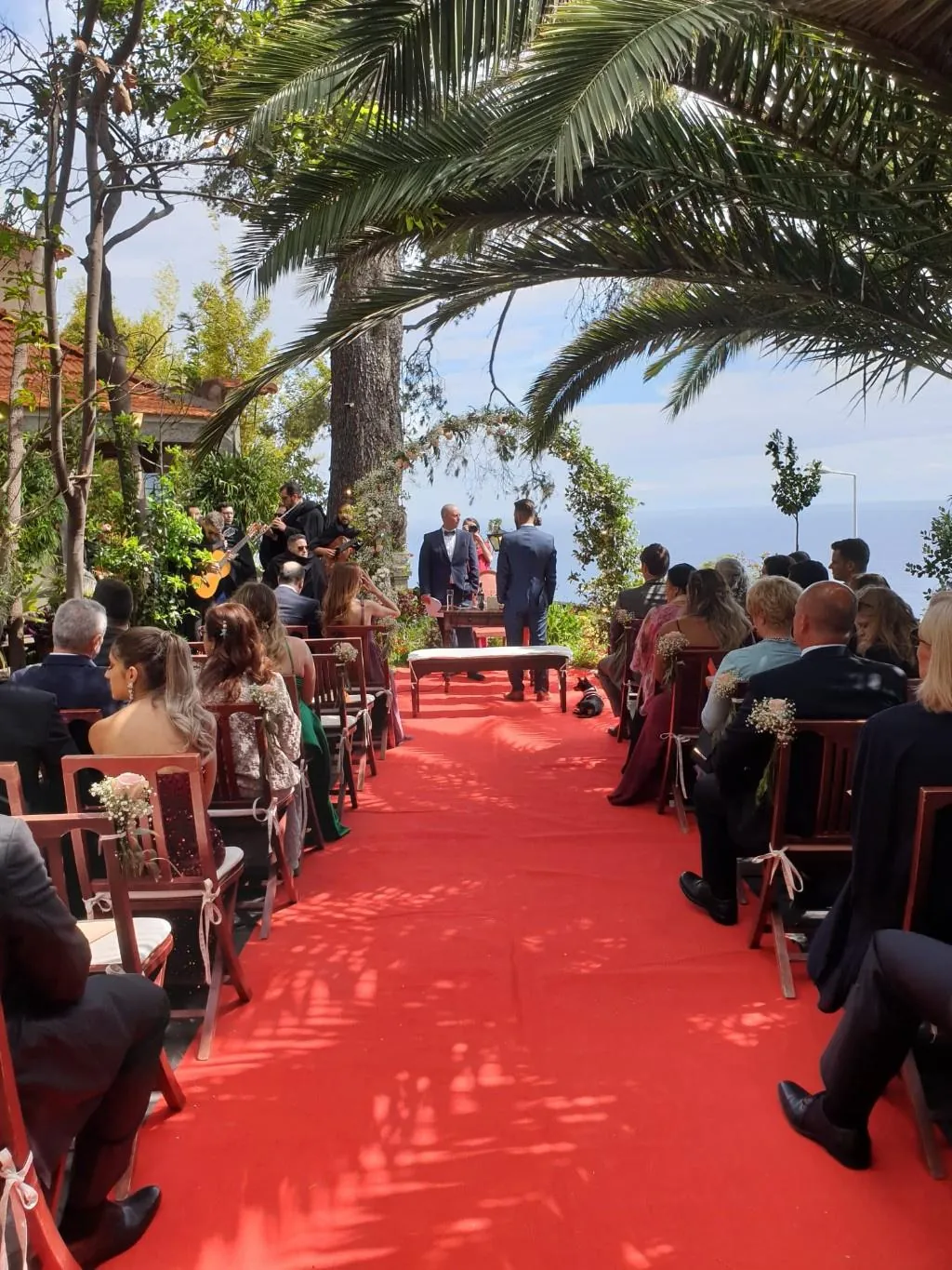 Cerimónia no jardim, com convidados dos dois lados da passadeira vermelha e o mar ao fundo