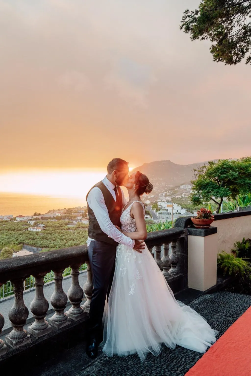 Noivos a beijar-se junto à balaustrada ao pôr do sol, com a encosta do Funchal ao fundo